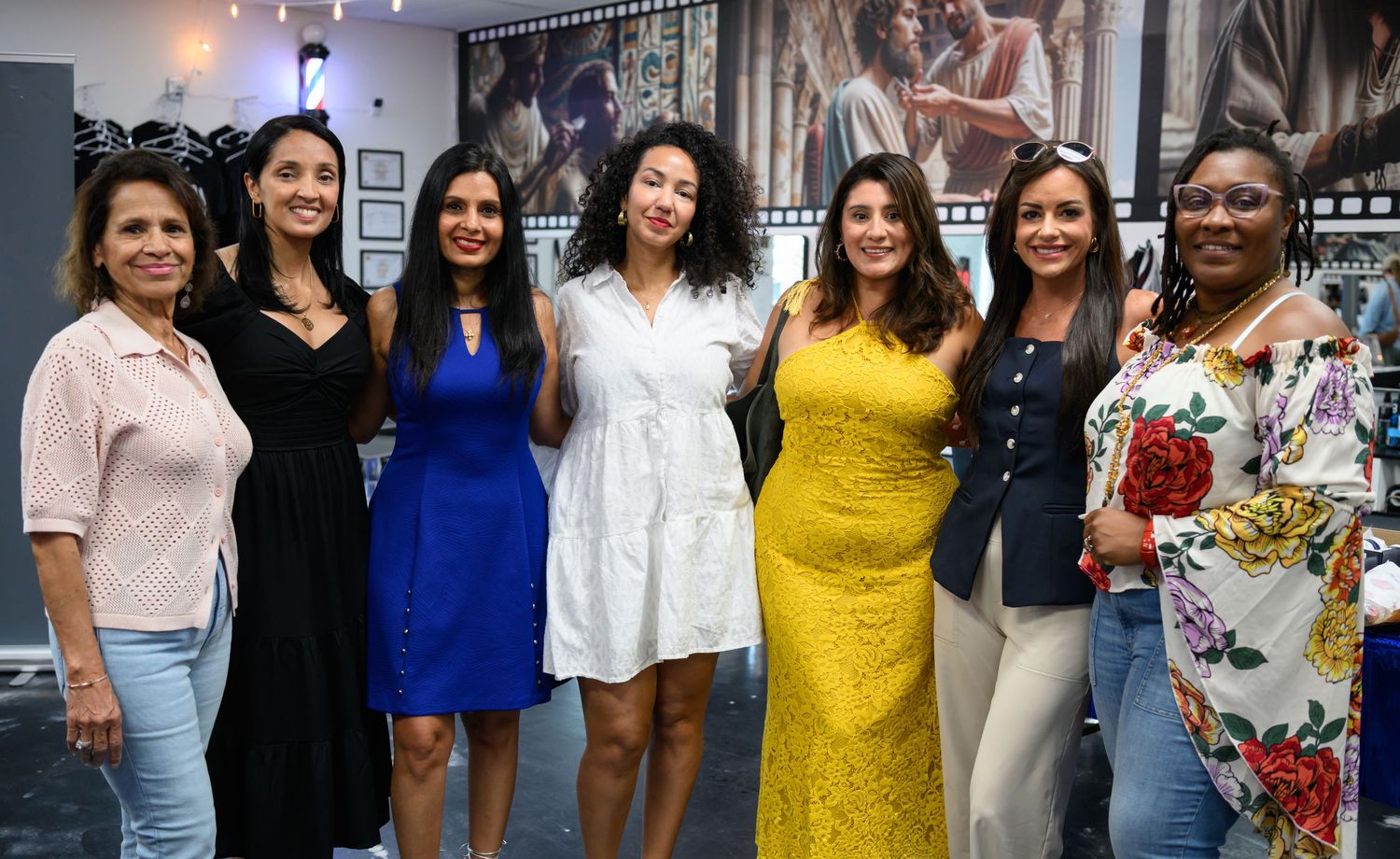 Seven women posing together smiling at an indoor event, wearing colorful dresses in a photography studio setting.