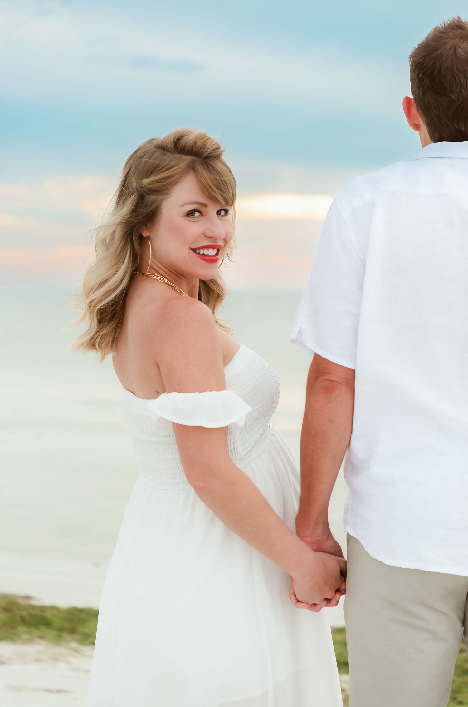 Pregnant woman in white off-shoulder dress holding hands with partner on beach, smiling over shoulder.