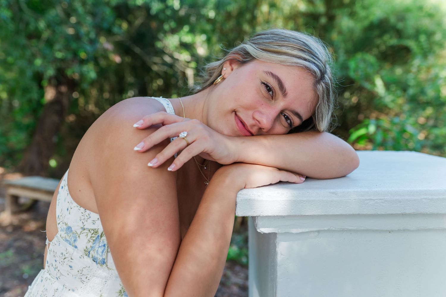 A smiling blonde woman in a floral dress leans on a white surface outdoors, showcasing an elegant ring on her hand.