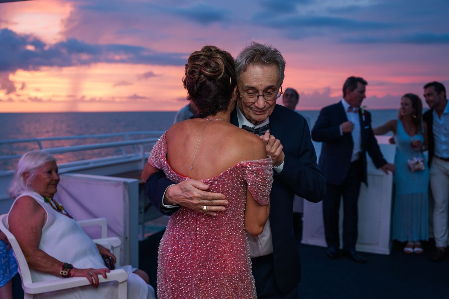 Couple dancing at sunset wedding reception on cruise ship deck with pink and purple sky over ocean.
