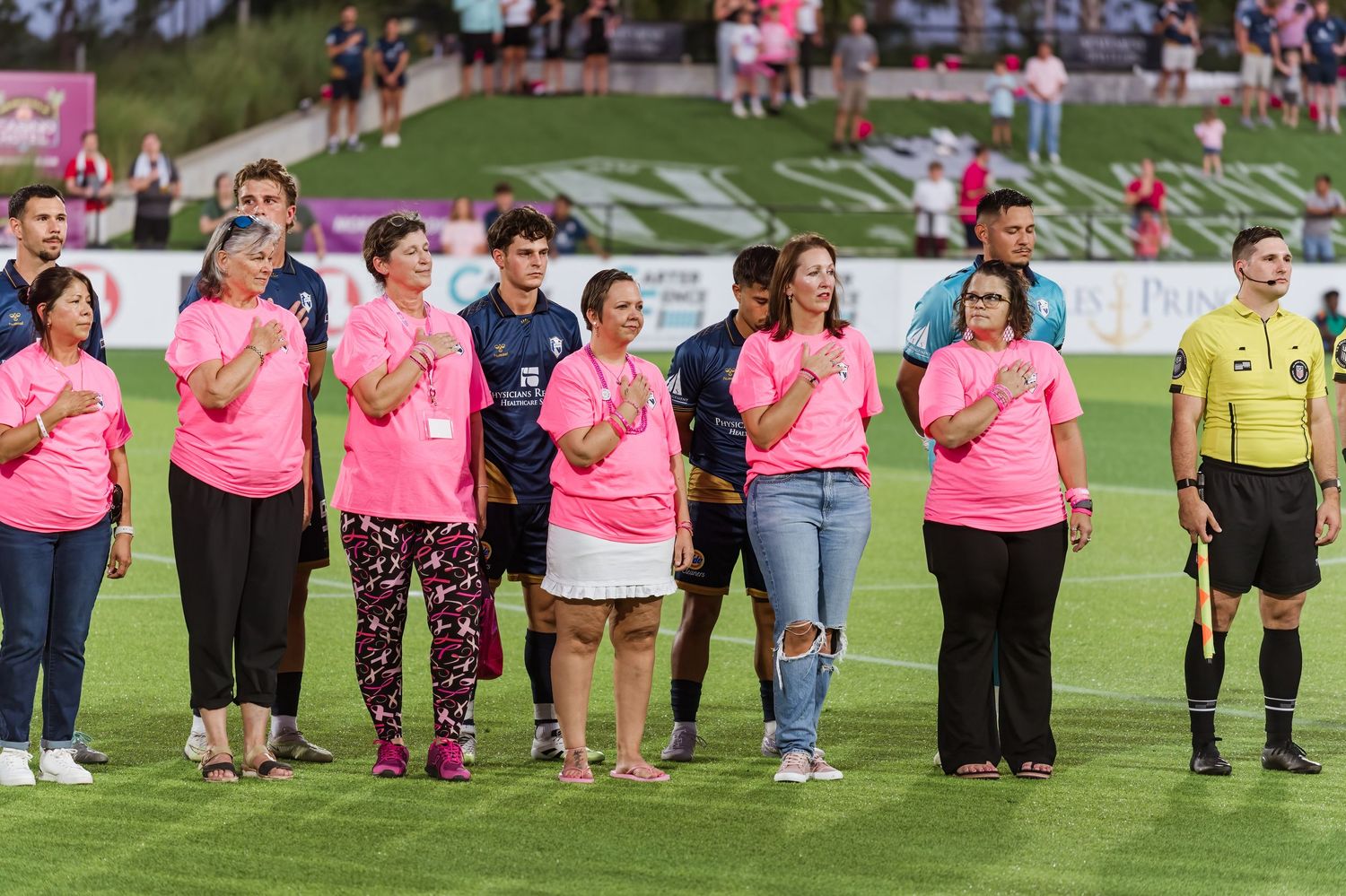 Soccer players and officials line up on a green field wearing pink and yellow uniforms during a pre-match ceremony.