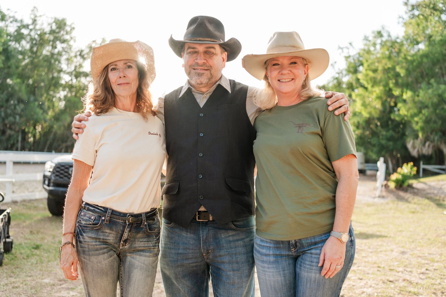 Three people in western attire pose together outdoors on a sunny day with trees in the background.