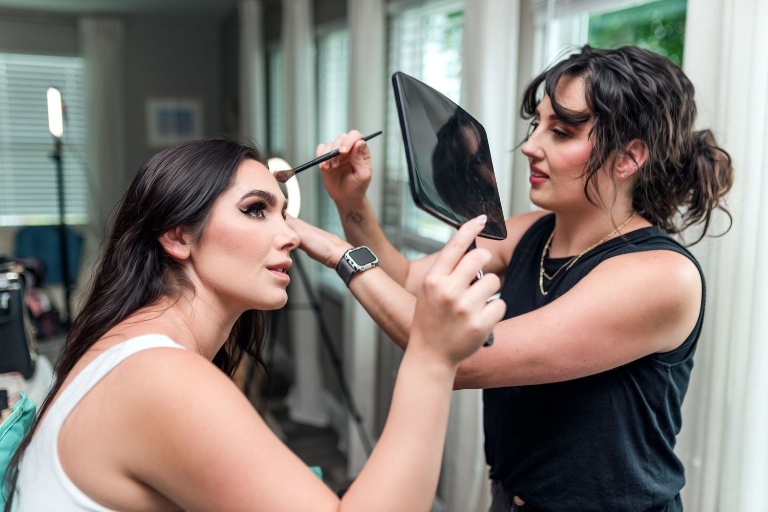 Four images show someone preparing makeup in a mirrored dressing room with professional lighting.
