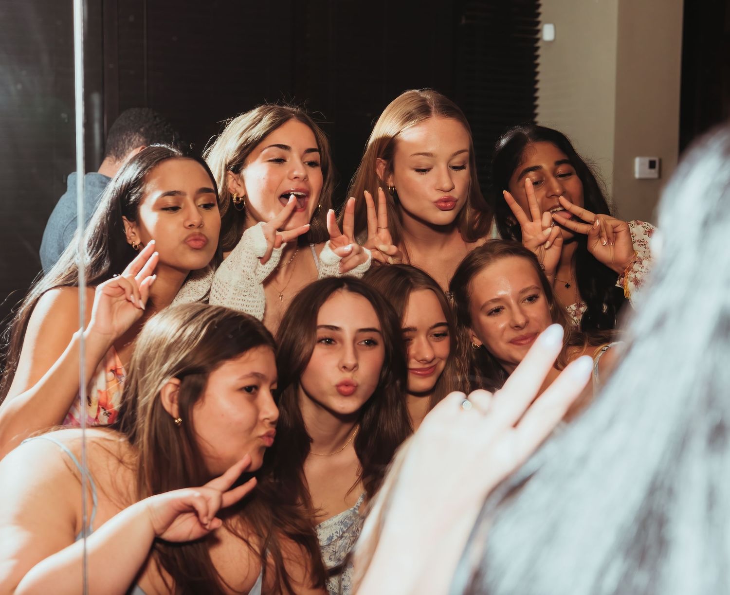 A group of bridesmaids gathered around the bride, blowing kisses and making playful poses for the camera in warm lighting.