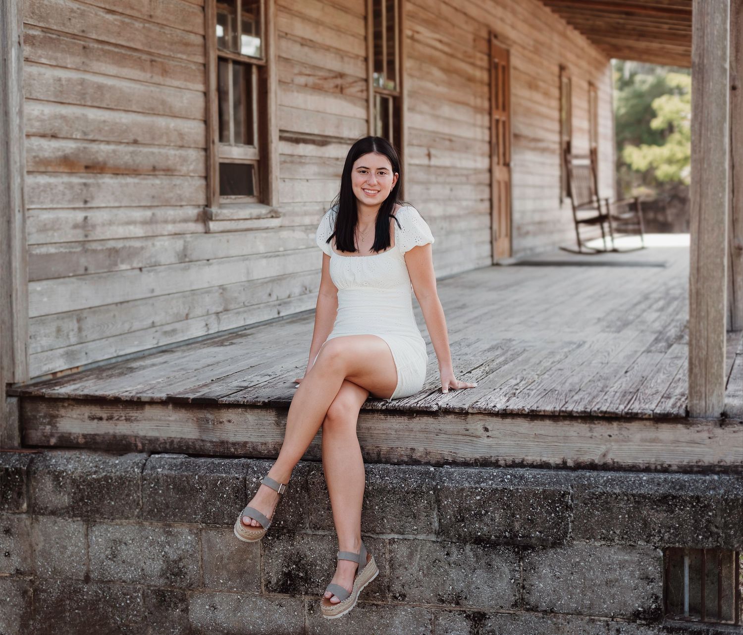 Woman in a white dress sits casually on a rustic wooden porch with rocking chairs.
