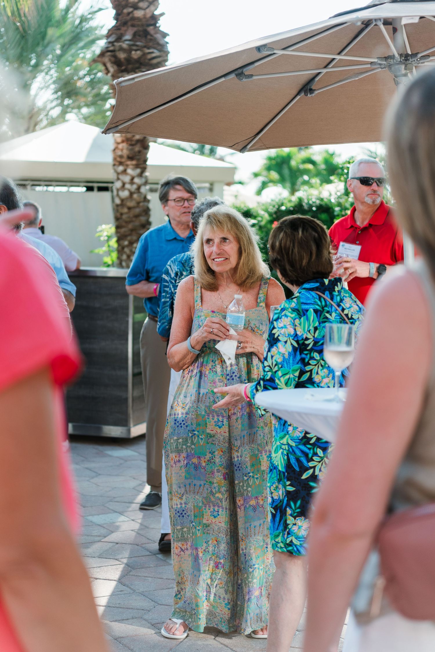 Woman in a colorful maxi dress smiling at an outdoor social gathering under beige umbrellas.