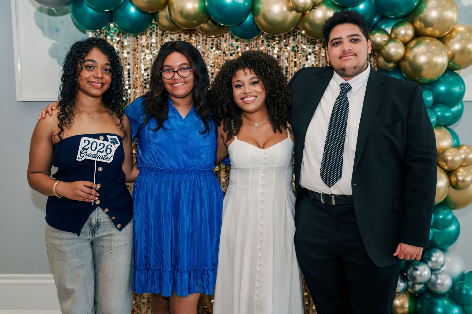 Four friends pose together at a graduation party with teal and gold balloon backdrop, one holding a 2020 grad sign.