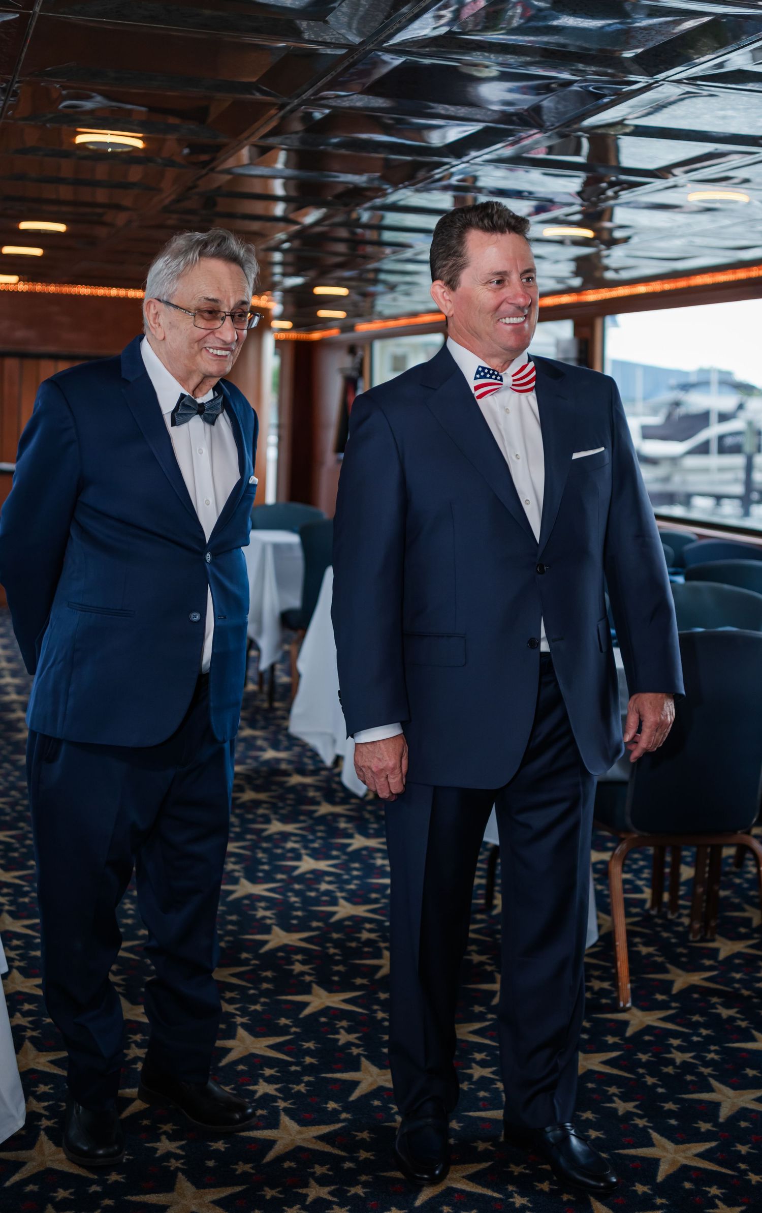 Two men in navy suits with patriotic bow ties smile together in an elegant cruise ship dining room.