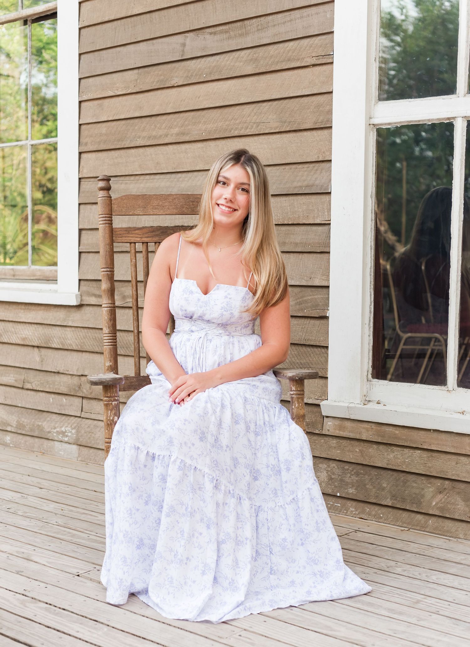 Someone in a white lace maxi dress sits on a wooden rocking chair on a rustic farmhouse porch.