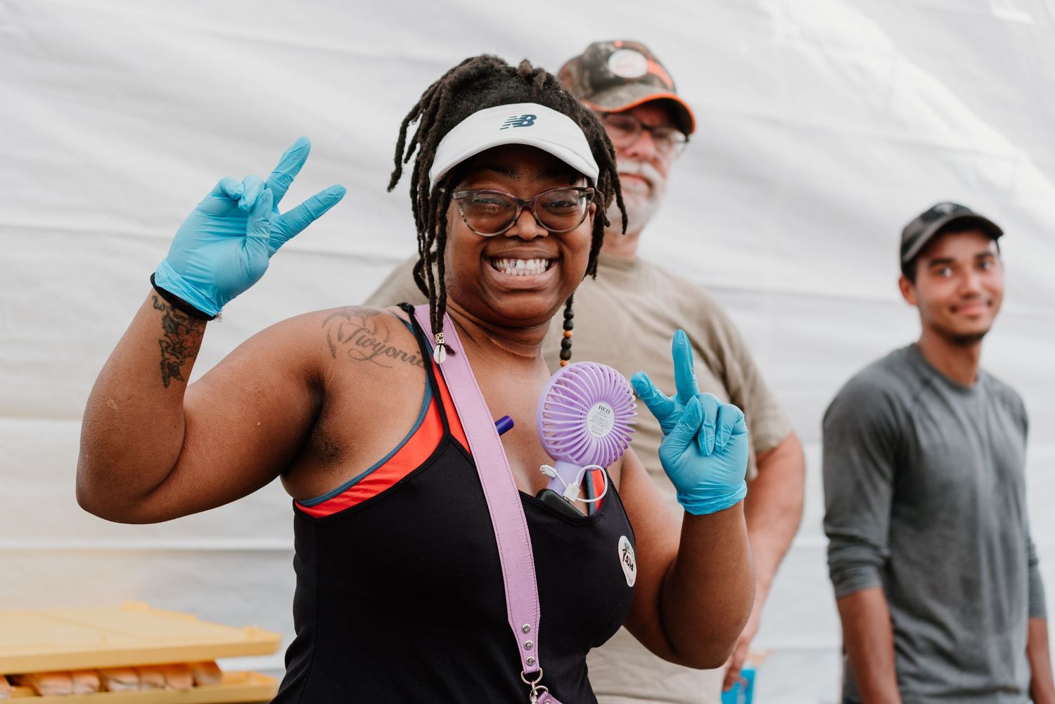 Happy volunteer wearing gloves makes peace sign gestures while outdoor with other team members.