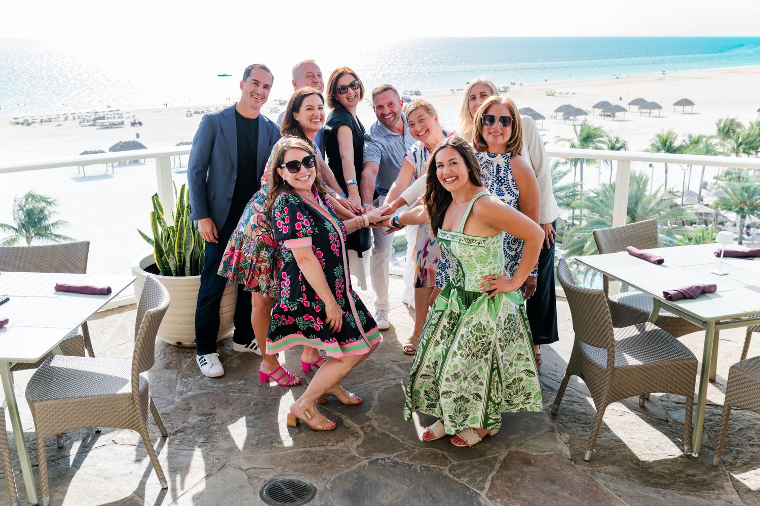 Group of smiling professionals posing together at an oceanfront restaurant terrace during a sunny day.