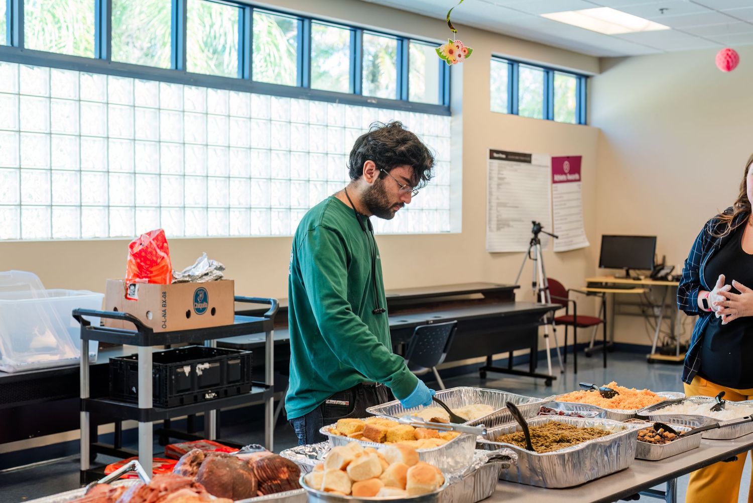 Students work on laptops and study materials in a bright classroom with large windows and fluorescent lighting.