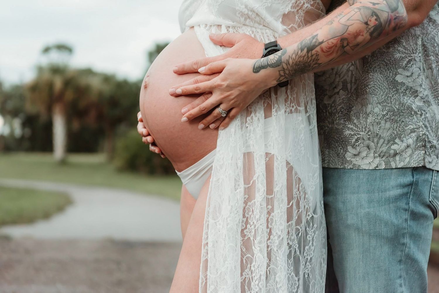 Person wearing white lace dress holds pregnant belly while standing outdoors with palm trees in background.