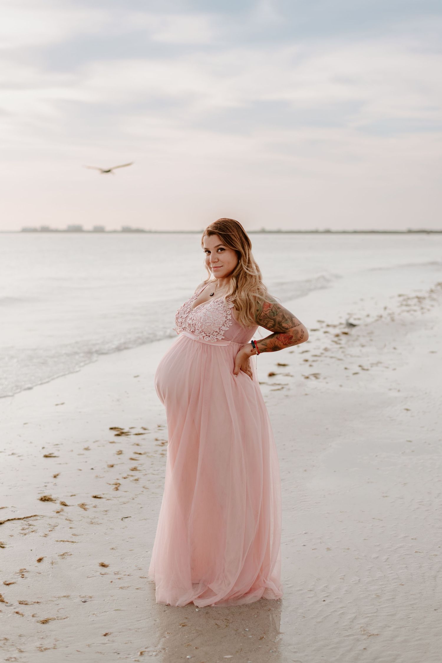A pregnant woman in a flowing pink dress stands gracefully on a serene beach at sunset with a bird flying overhead.