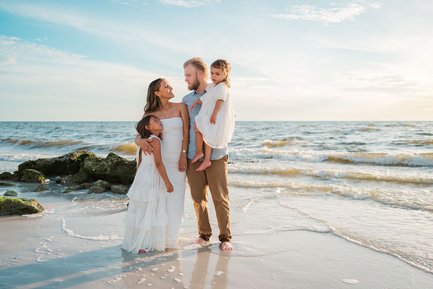 Family of four posing on a sandy beach at sunset, waves gently washing ashore near rocky coastline.