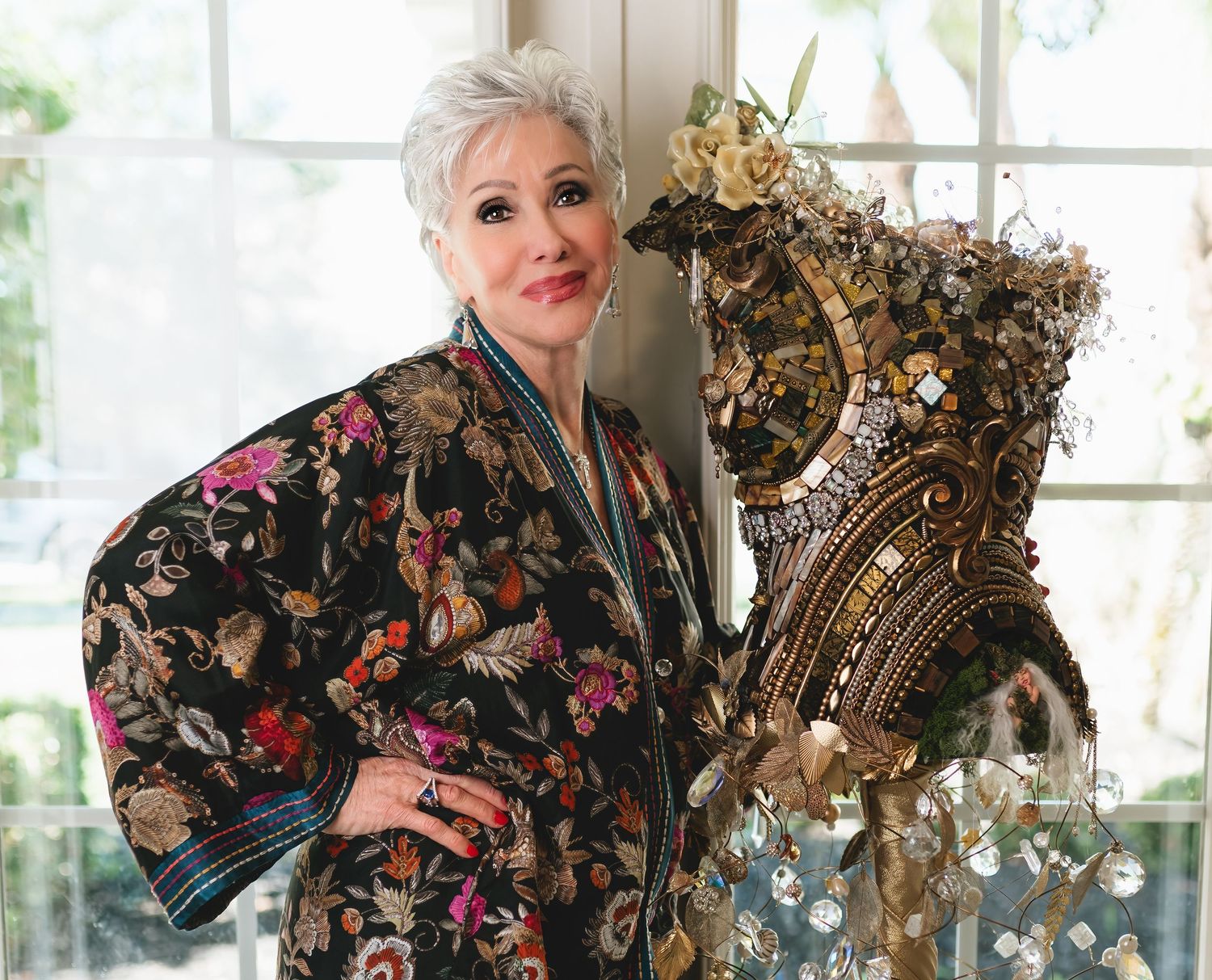 Sophisticated woman in floral kimono poses beside an ornate jeweled mannequin torso in a bright studio.