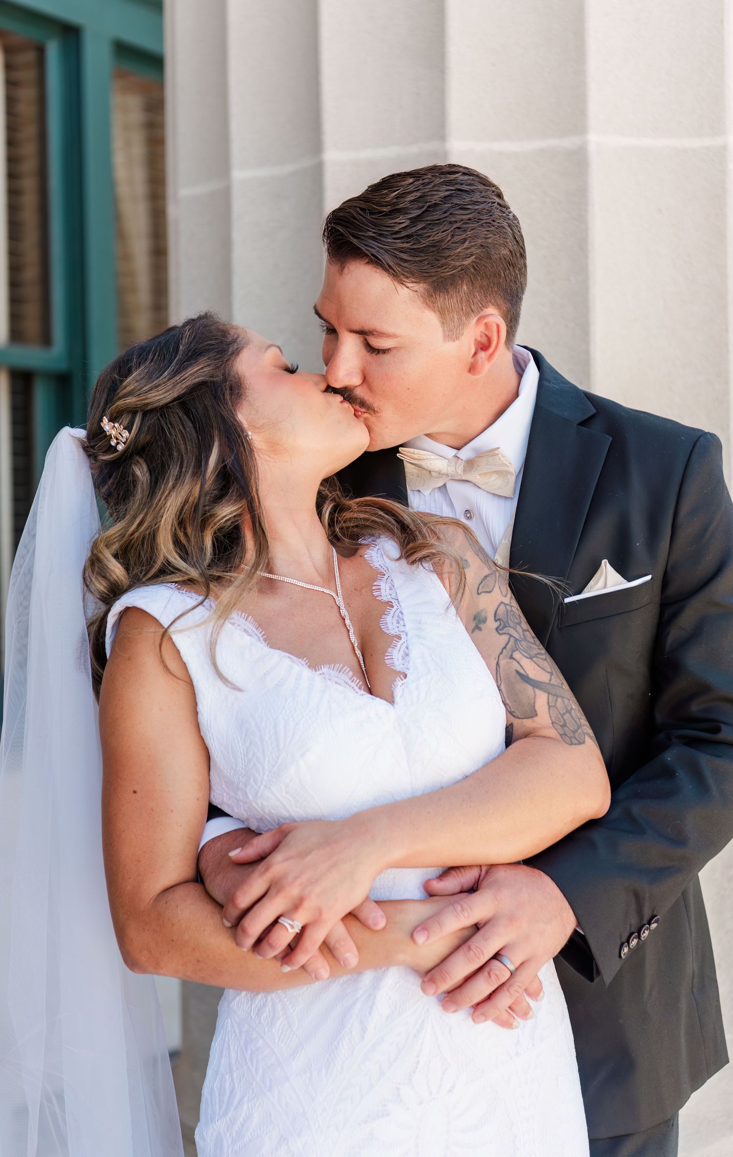 Bride and groom share romantic kiss outdoors, bride in lace gown with veil, groom in navy suit.
