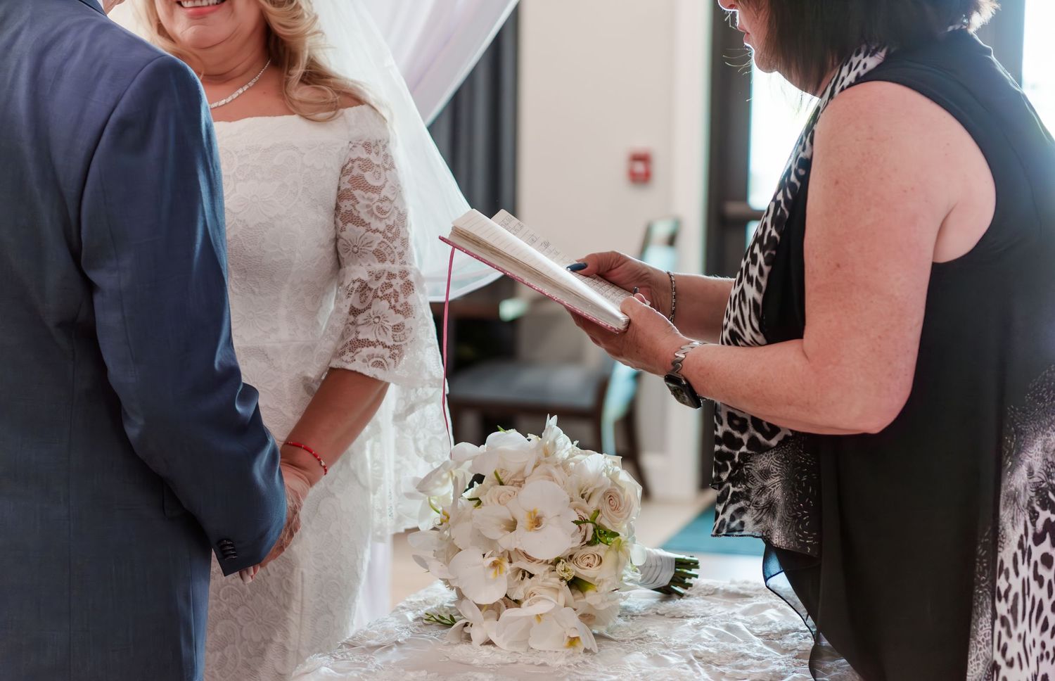 A bridal bouquet of white flowers rests on a table during a wedding ceremony indoors.