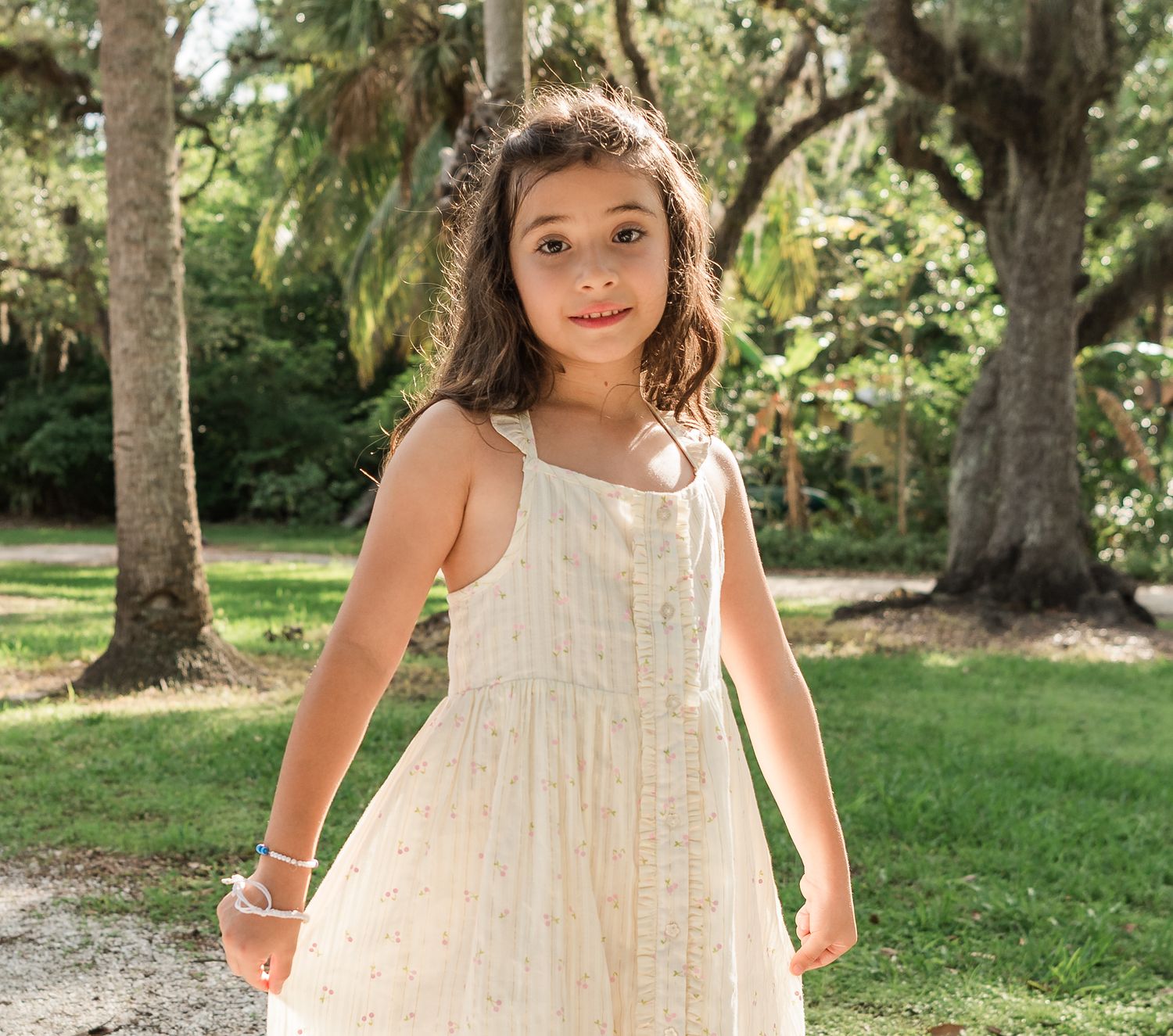 A child in a flowing white lace dress twirls and plays in a serene garden setting under the shade of mature trees.