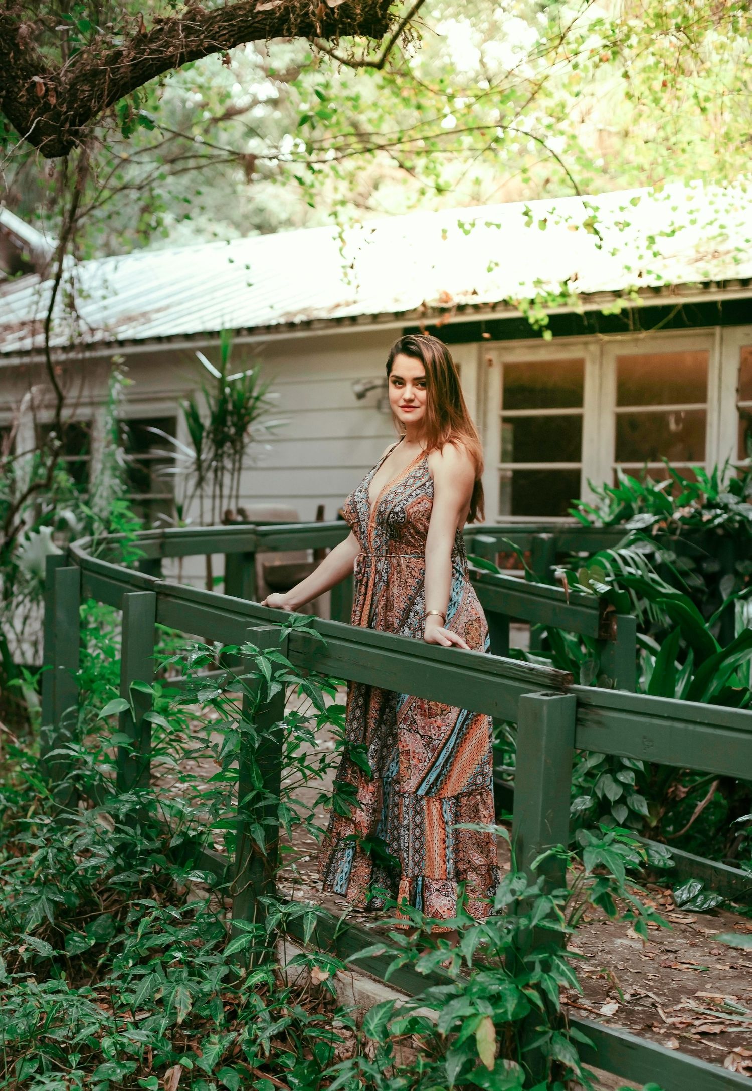 A person in a floral dress poses gracefully on green wooden stairs surrounded by lush garden foliage.