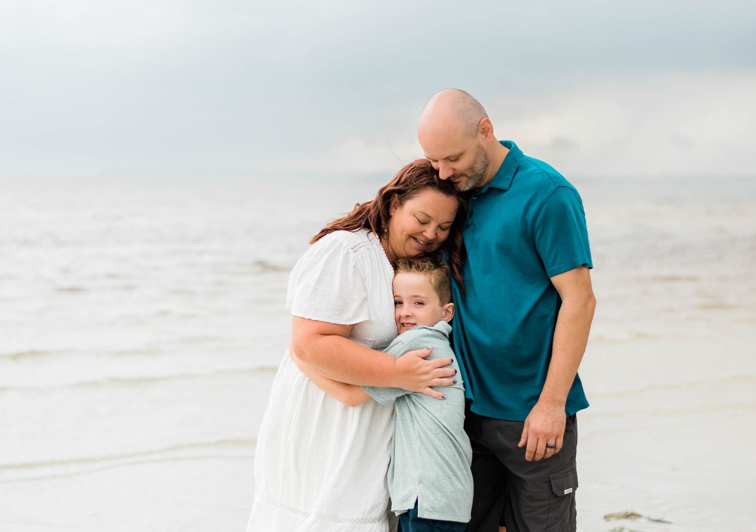 Family stands close together on a sandy beach in soft muted colors during sunset.