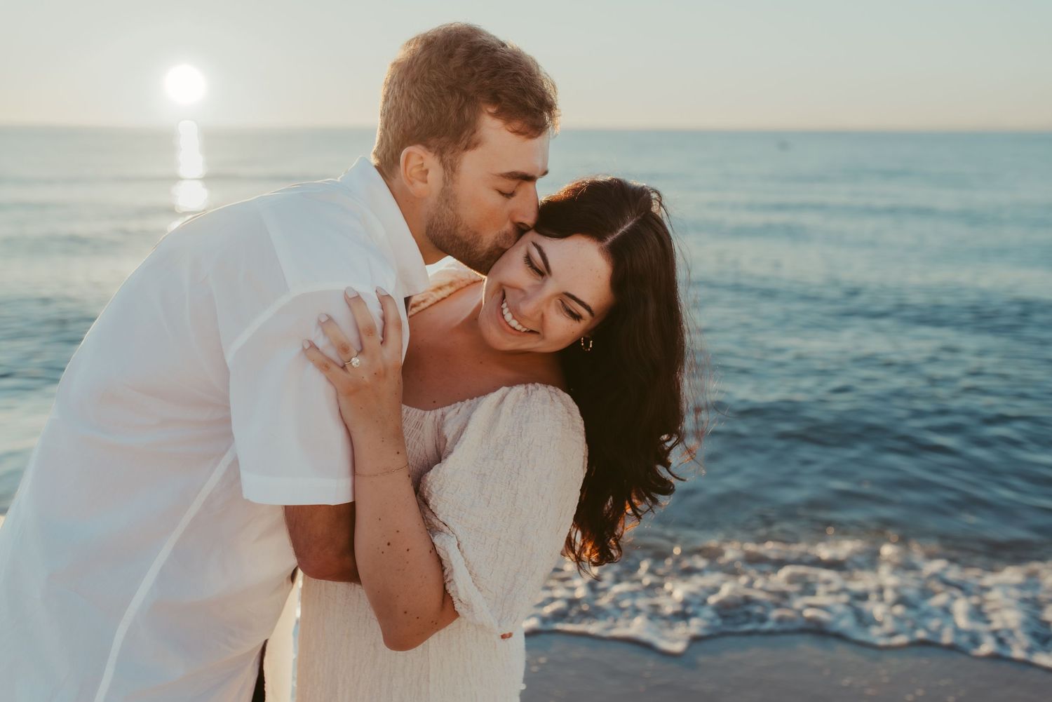 A romantic couple embraces lovingly by the ocean at sunset, with sunlight sparkling on the waves behind them.
