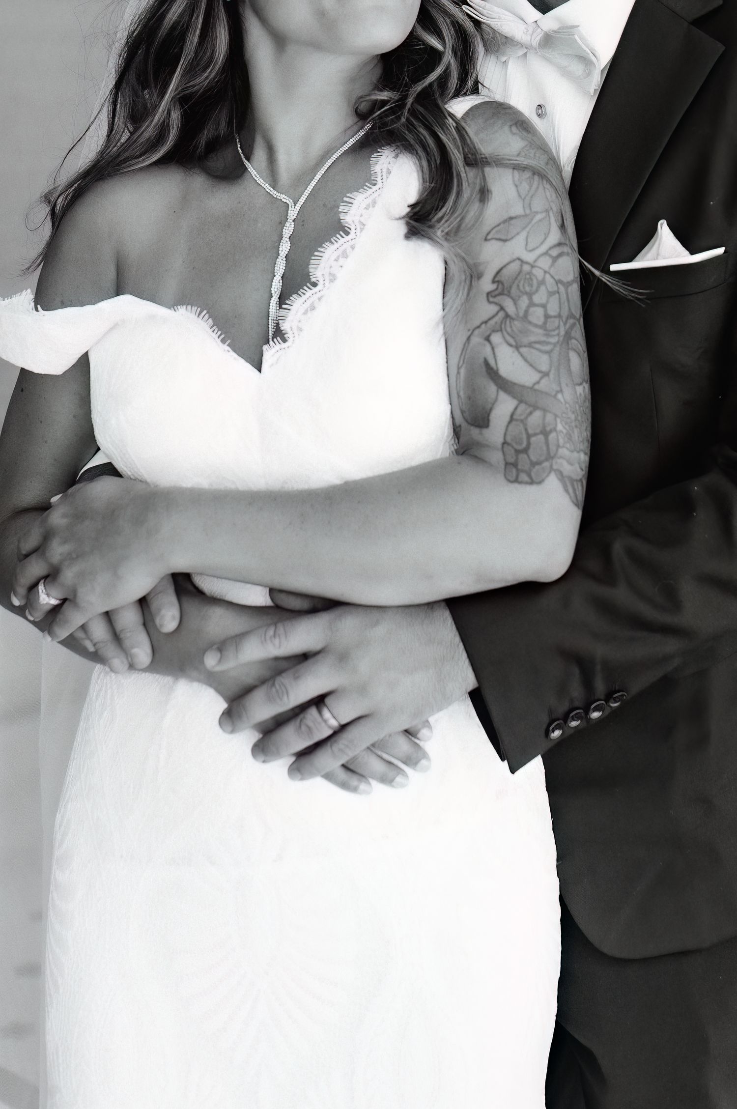 Black and white close-up of bride and groom embracing, showcasing wedding rings and bride's floral arm tattoo.