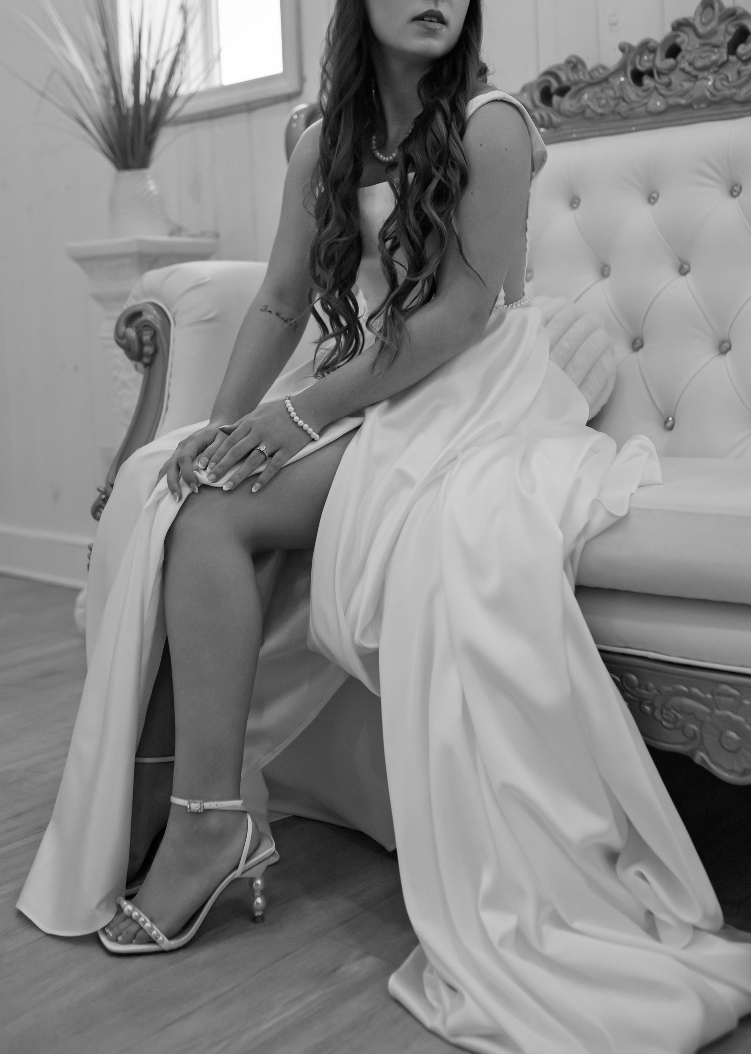 Black and white photo of a bride in a white gown with a slit, seated on a tufted sofa wearing strappy heels.