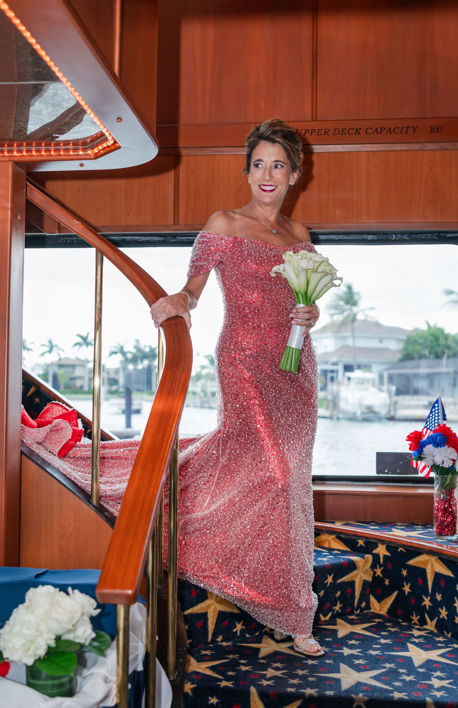 Woman in pink sequin gown holding white calla lilies bouquet on yacht staircase with patriotic decor.