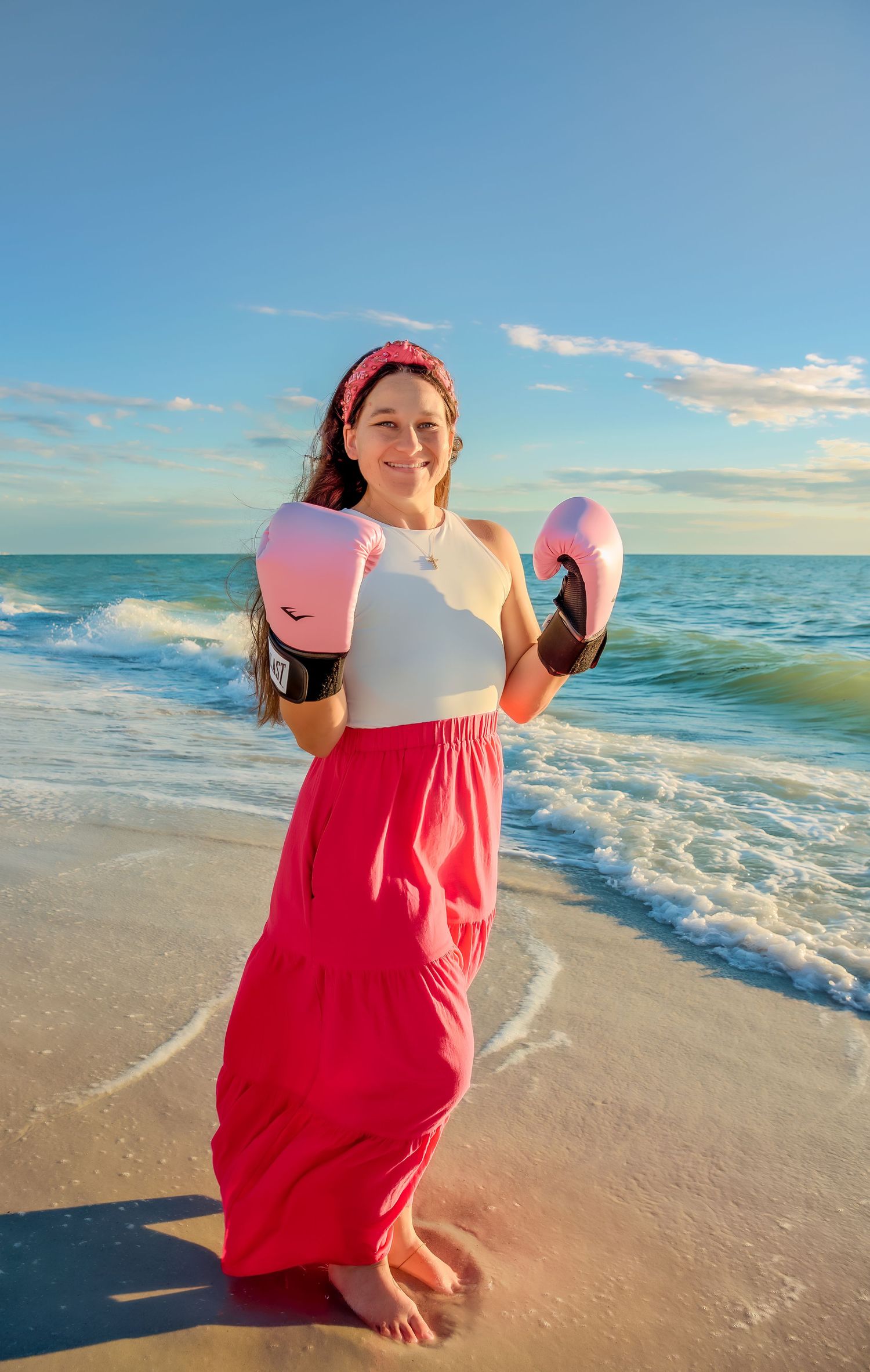 Person in a bright red dress holding a surfboard poses playfully on a sunny beach at sunset.