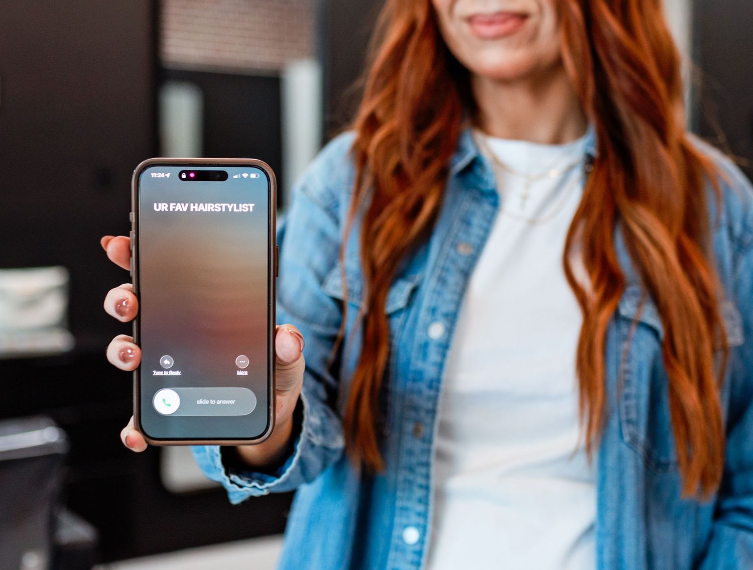 Person with red hair wearing denim jacket and white top uses smartphone while standing against dark background.