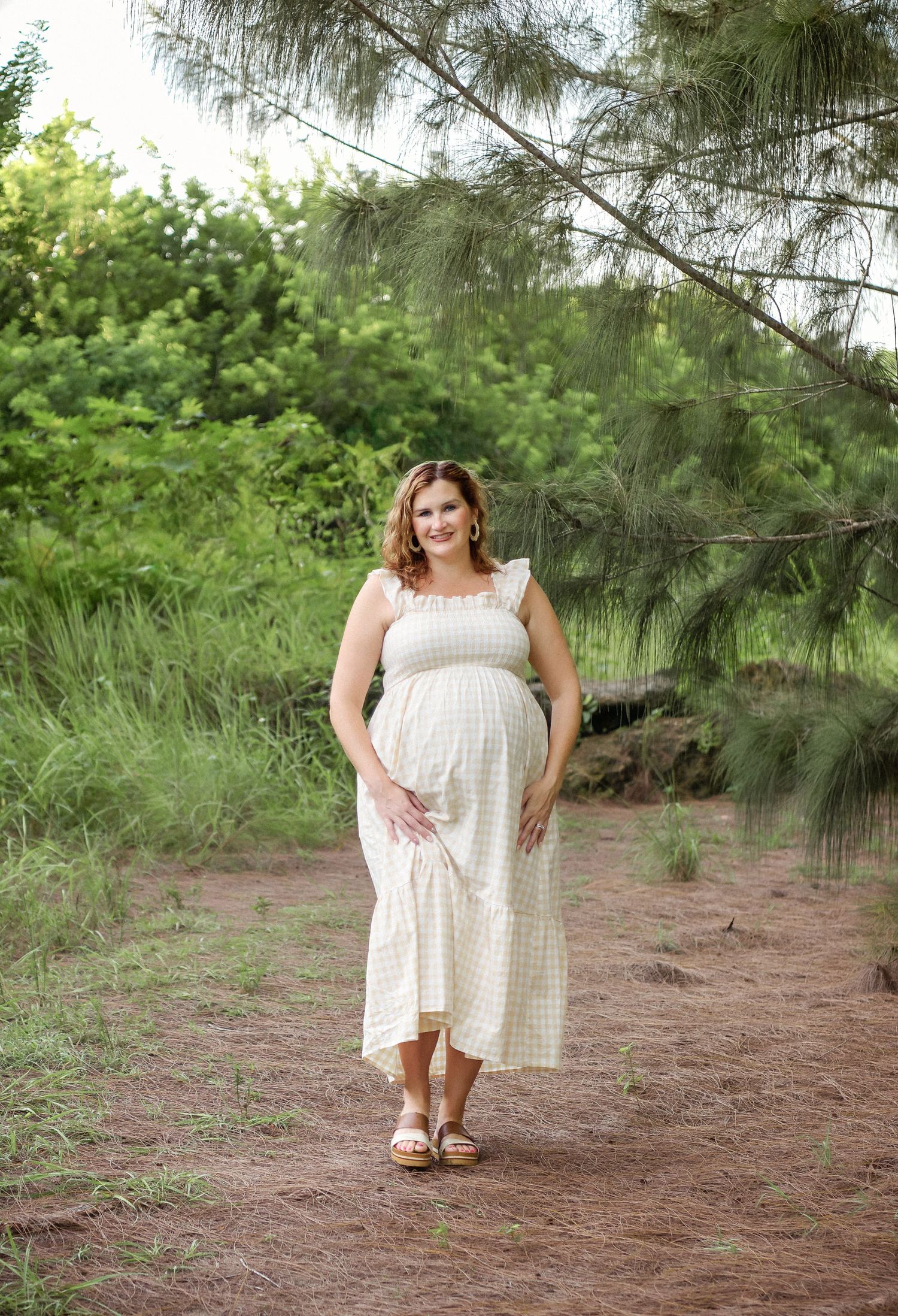 A woman in a white summer dress poses outdoors among pine trees on a dirt path in a natural setting.