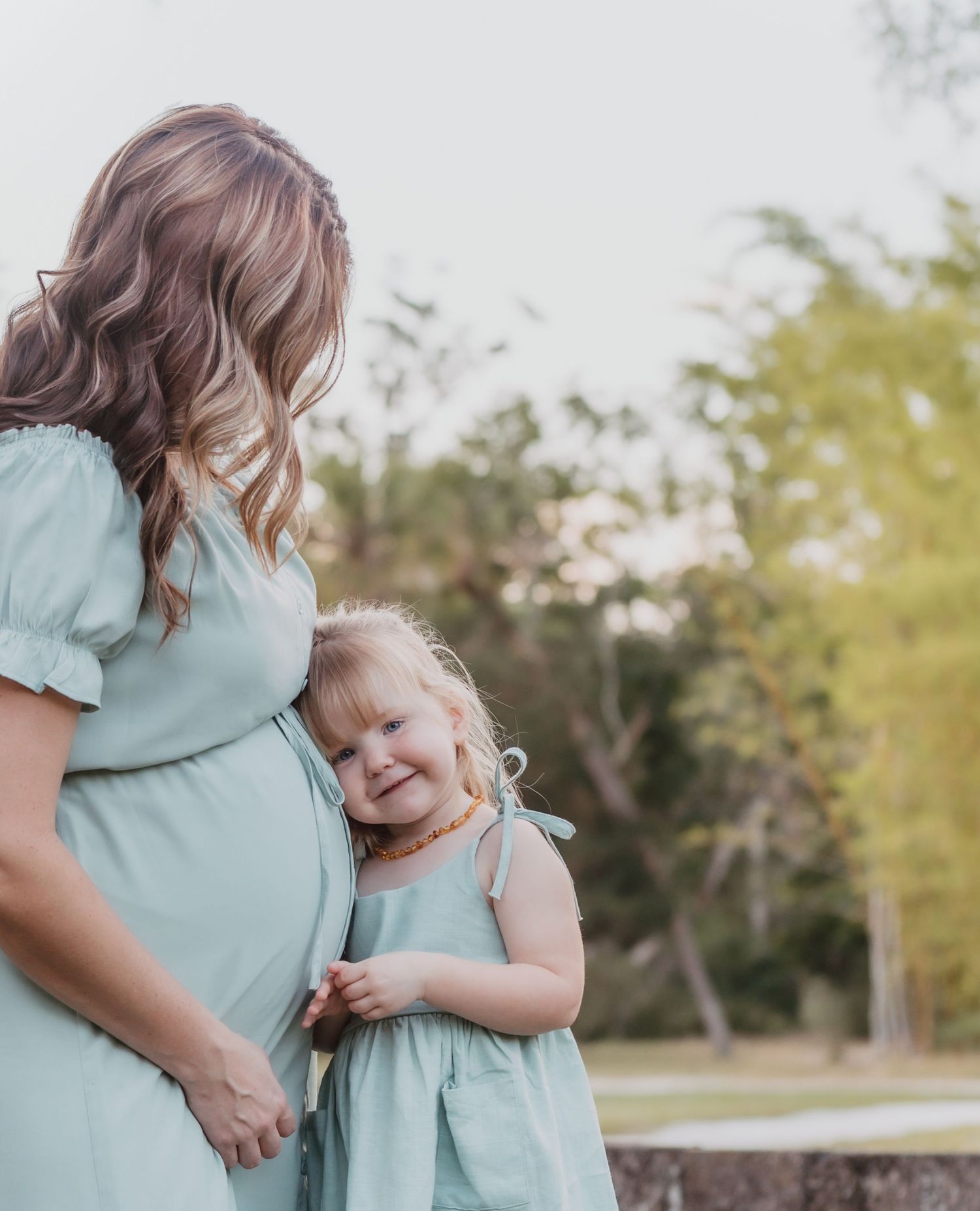 A mother in a mint green dress stands with her young daughter in matching outfits during a sunny outdoor photoshoot.