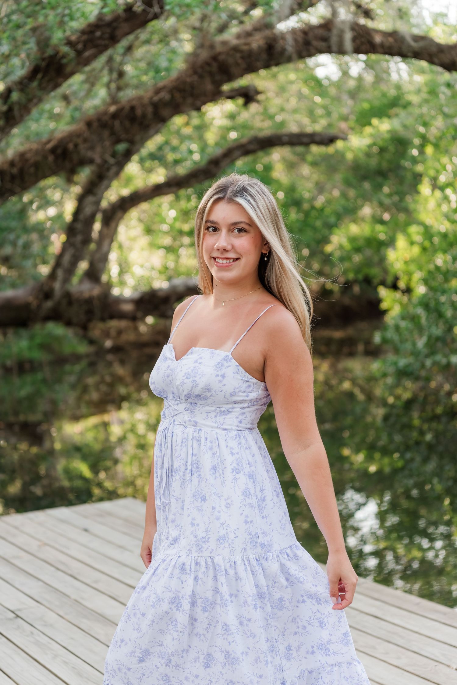 Woman in a white floral sundress stands on wooden deck with trees and water in background on sunny day.