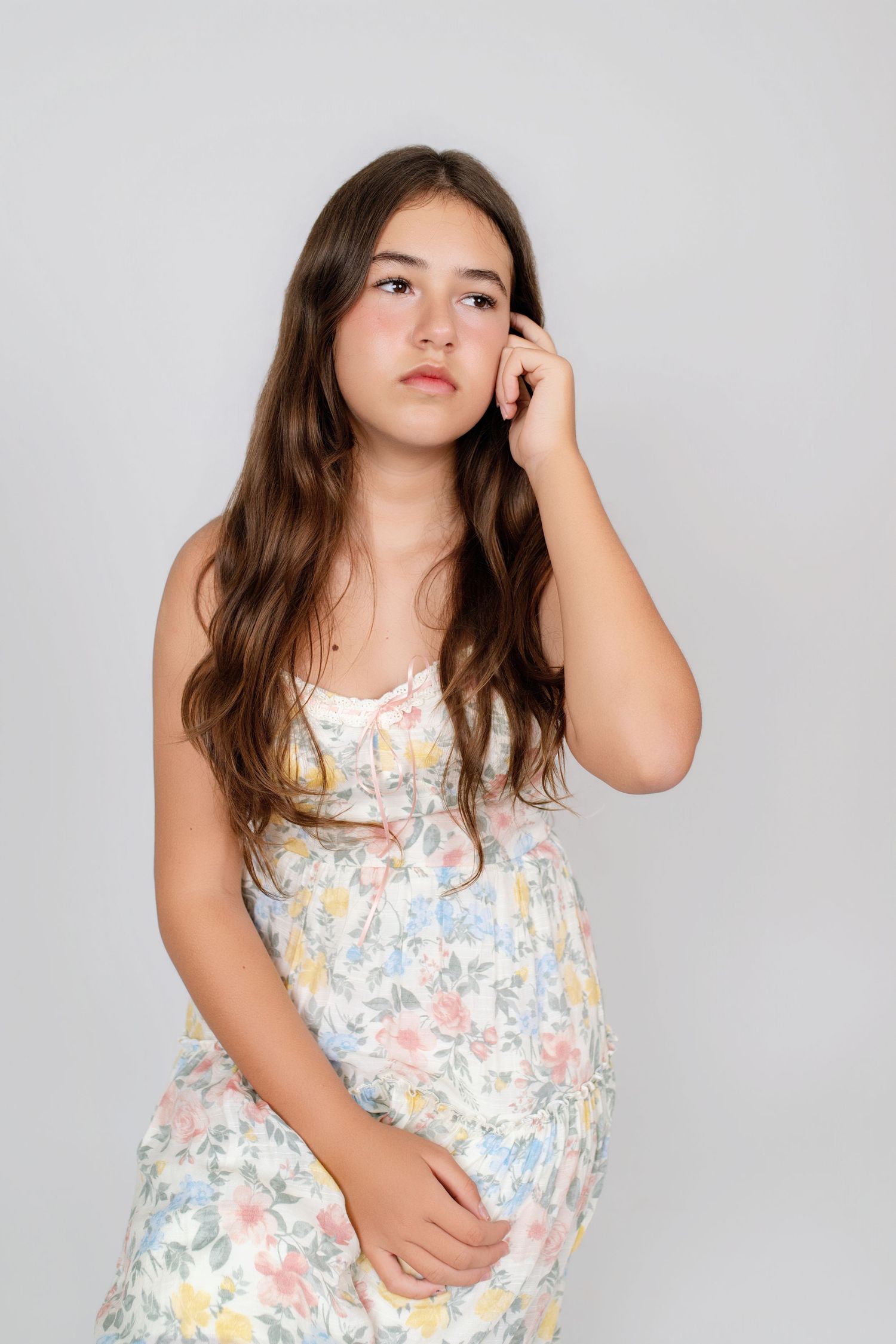 Young girl in floral sundress with long brown hair posing thoughtfully against a light gray background.