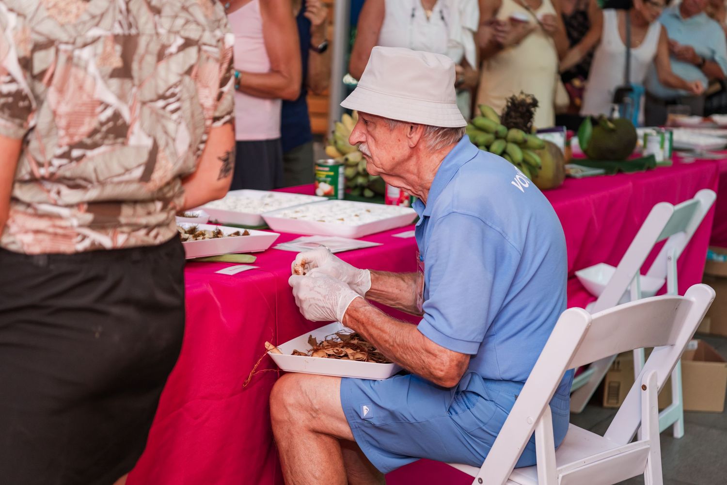 Elderly man in blue shirt and white hat eating at an outdoor food event with pink tablecloth.