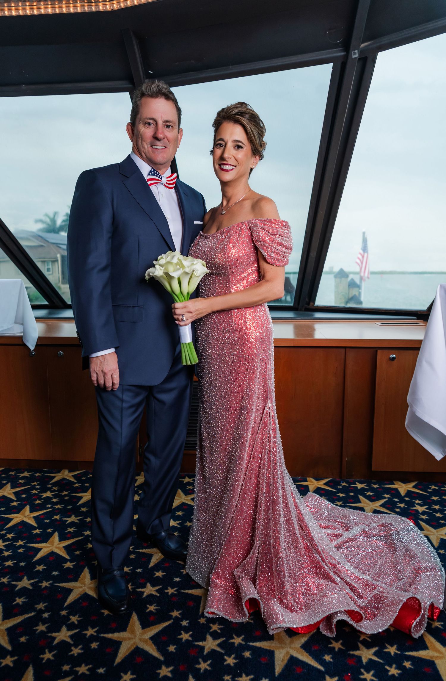 Elegant couple posing in formal attire, man in navy suit with bow tie, woman in pink sequin gown holding white bouquet.