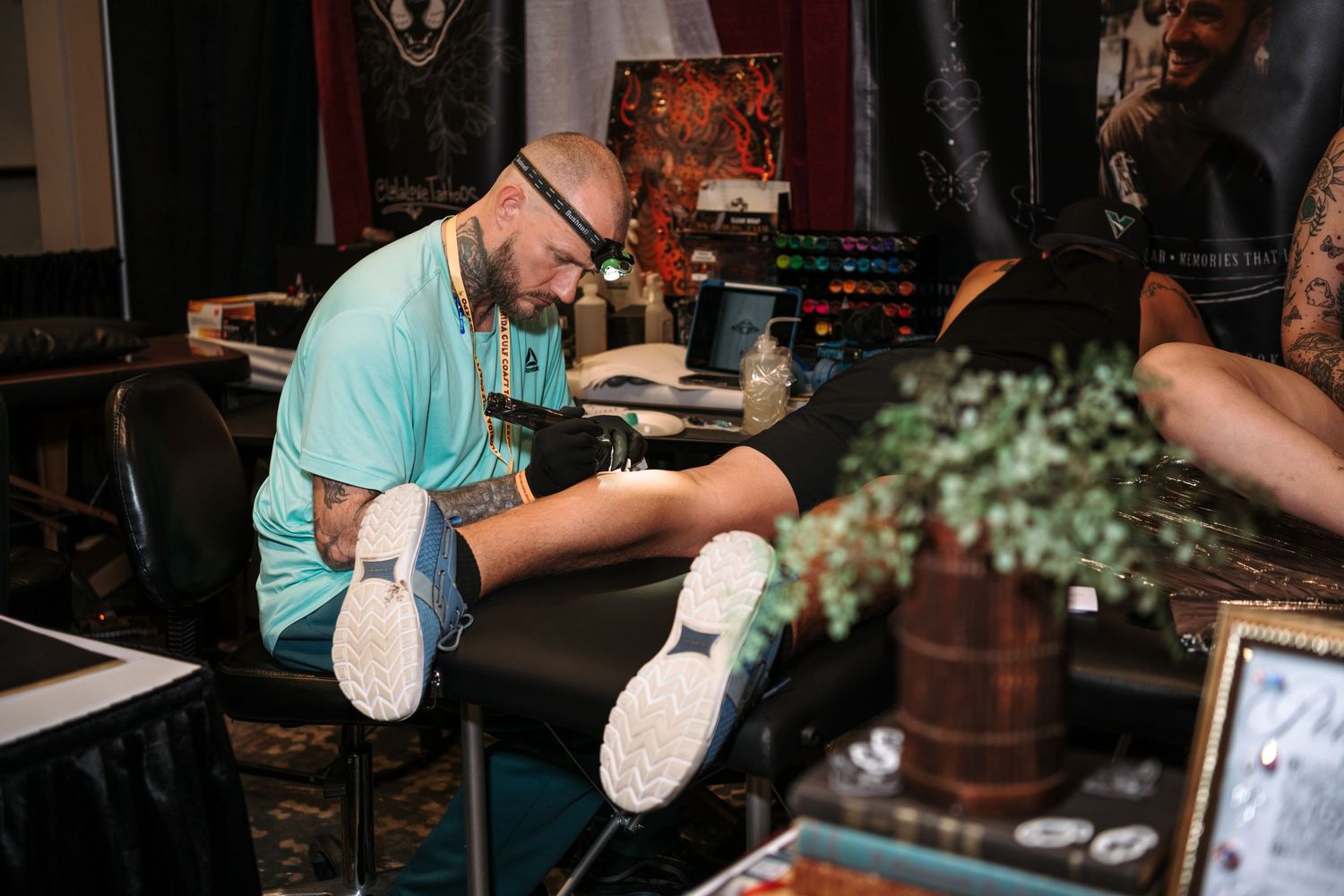 Tattoo artist in teal shirt applying tattoo on client's leg at a tattoo convention booth.