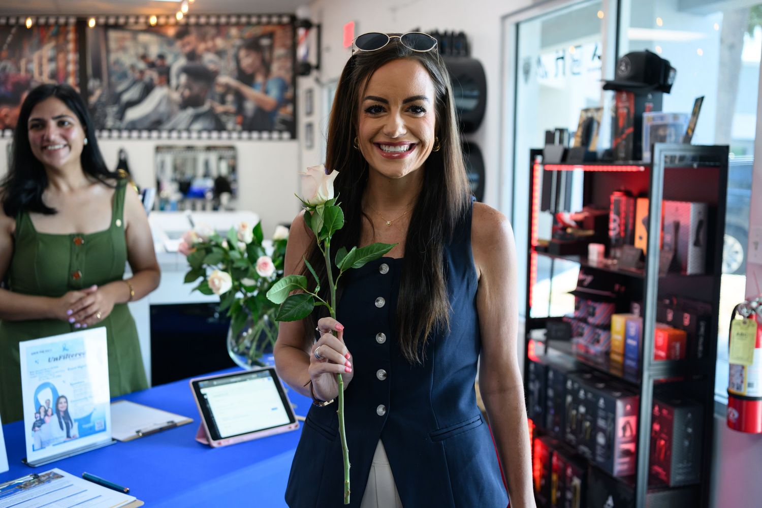 A smiling woman holds a white rose inside a boutique shop, with shelves of products and another woman visible behind her.