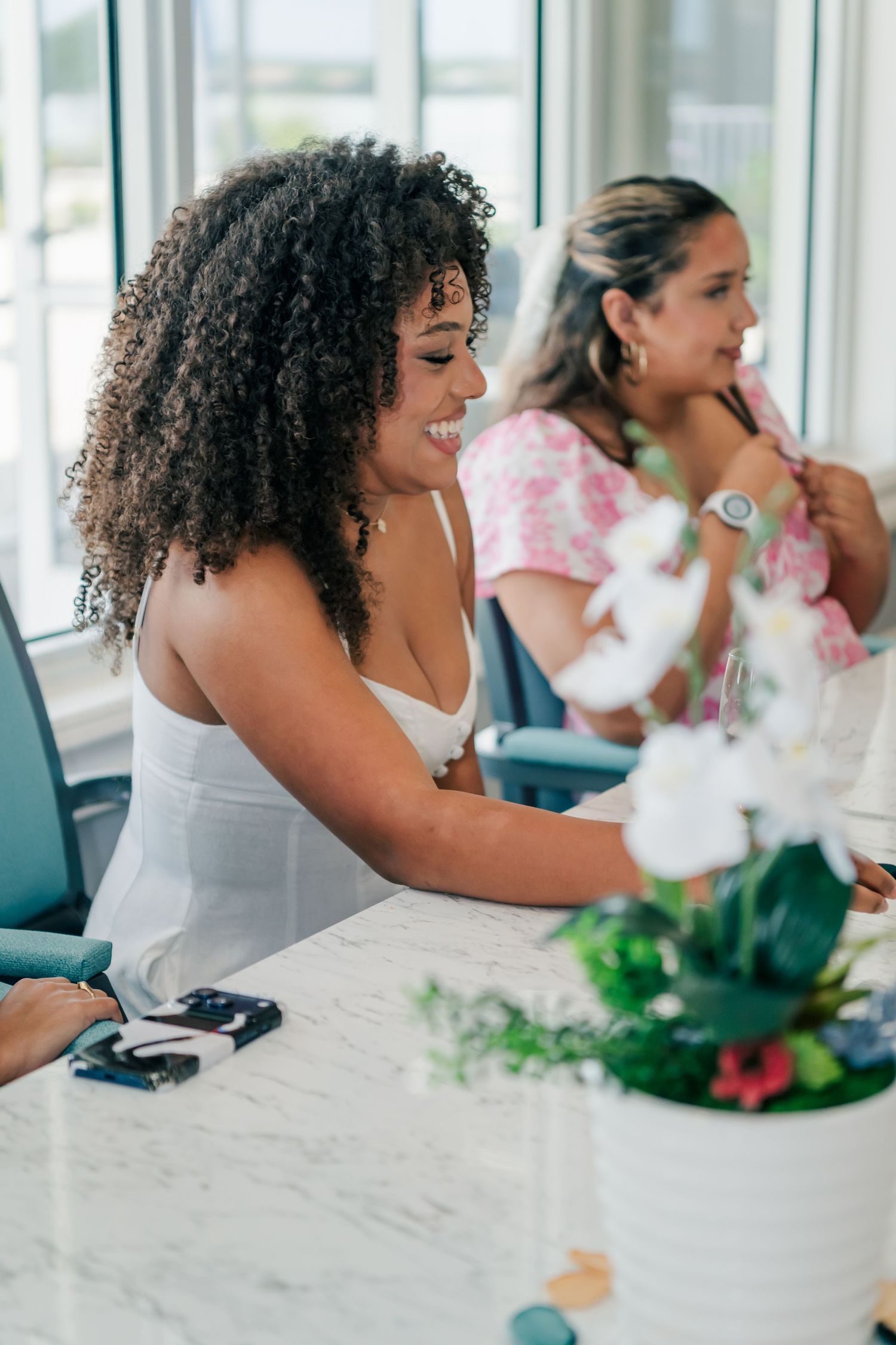Two women smiling and laughing together at a marble table with white orchids in a bright, airy restaurant setting.