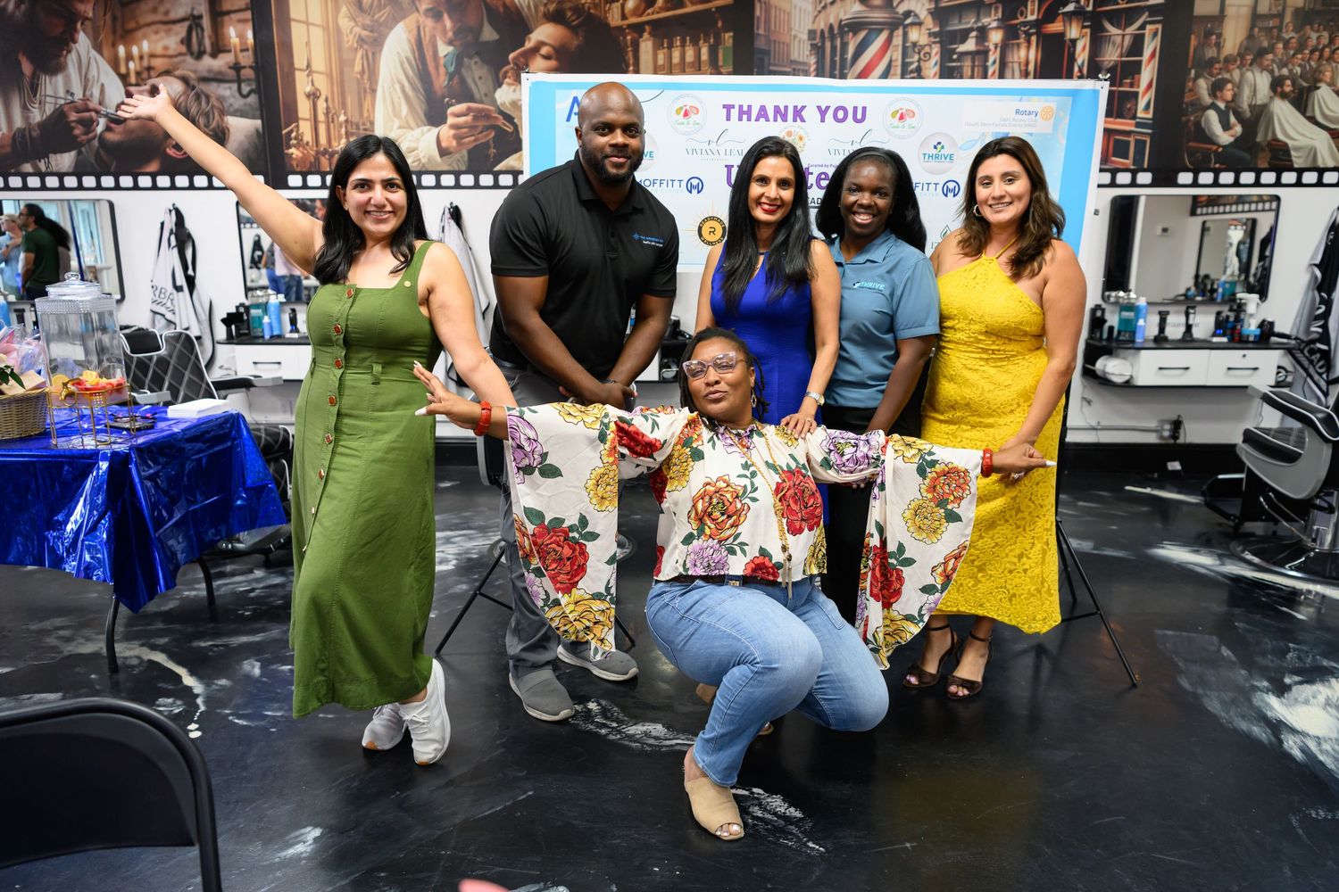 Group of six diverse people posing and smiling in front of a Thank You banner at an indoor event.
