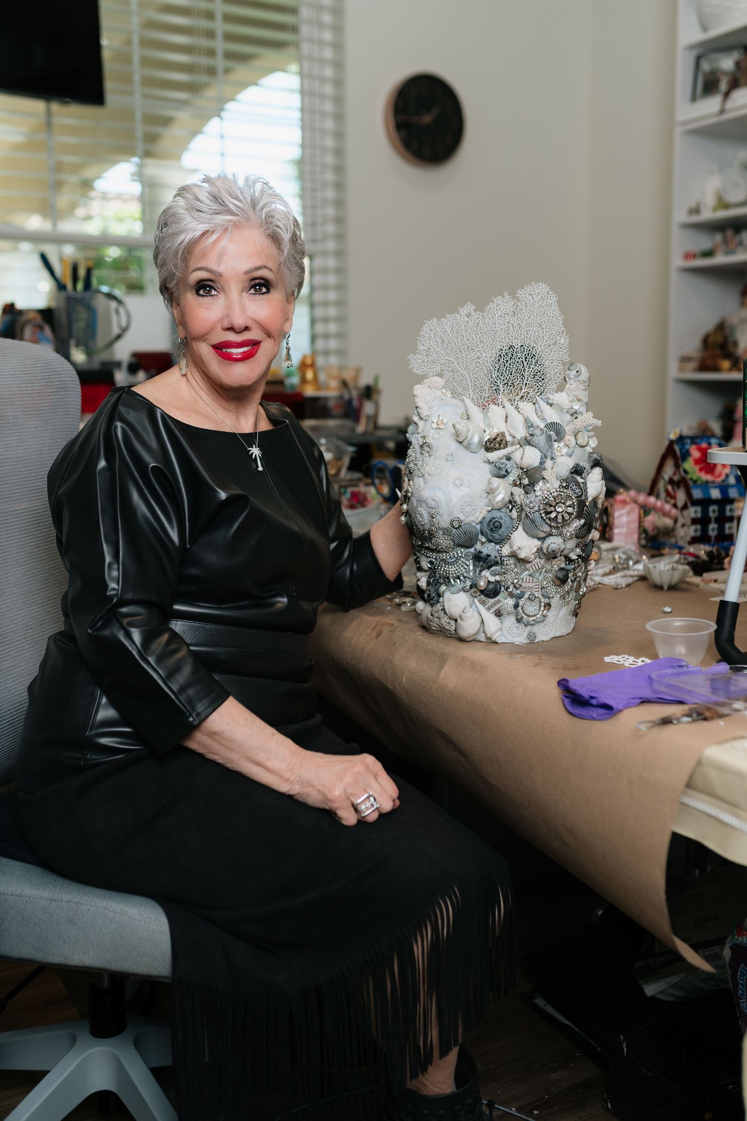 Elegant older woman in black dress poses beside an ornate shell and crystal decorative sculpture at her desk.