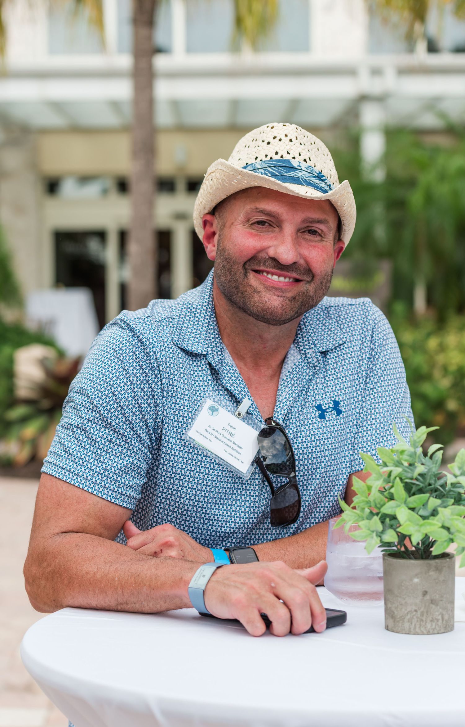 Smiling man in straw hat and blue polo shirt sitting at outdoor table with name badge and sunglasses.