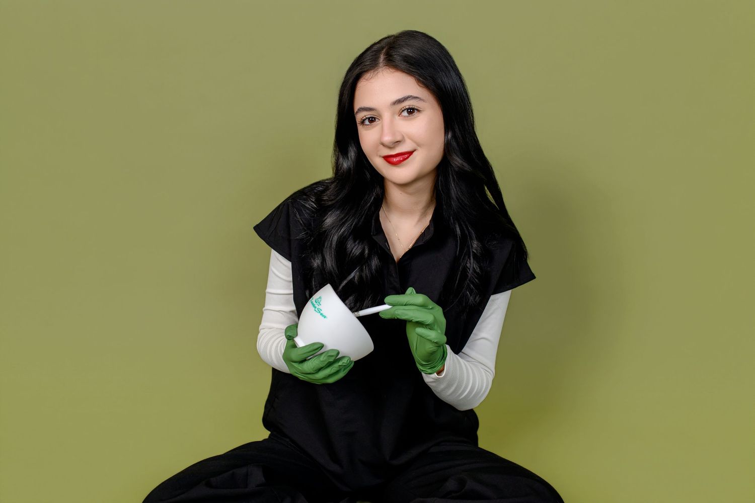 Young woman in black dress with green gloves holding a white bowl and spoon against olive background.