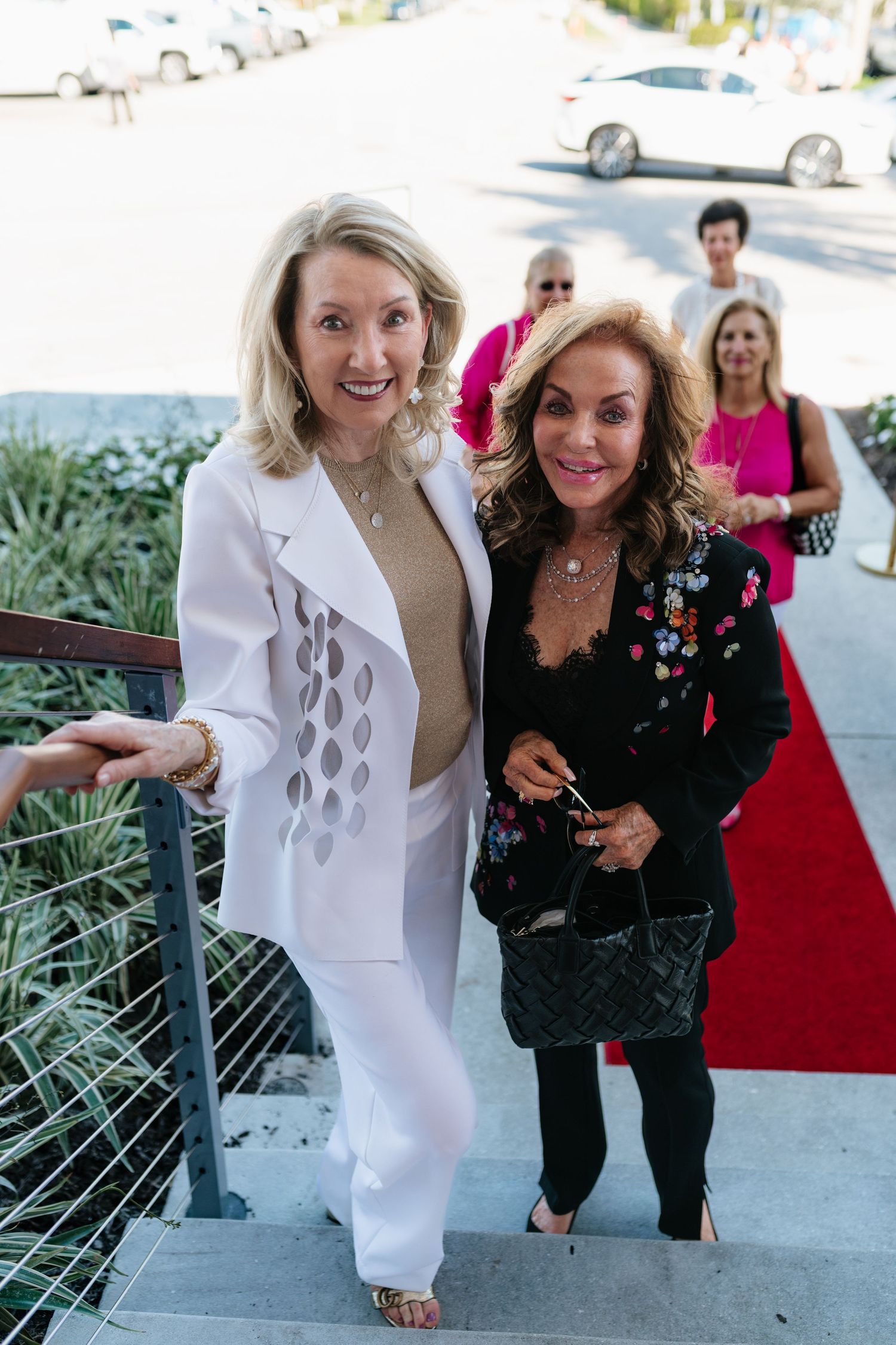 Two elegantly dressed women pose together on a red carpet with palm trees and cars visible in the background.