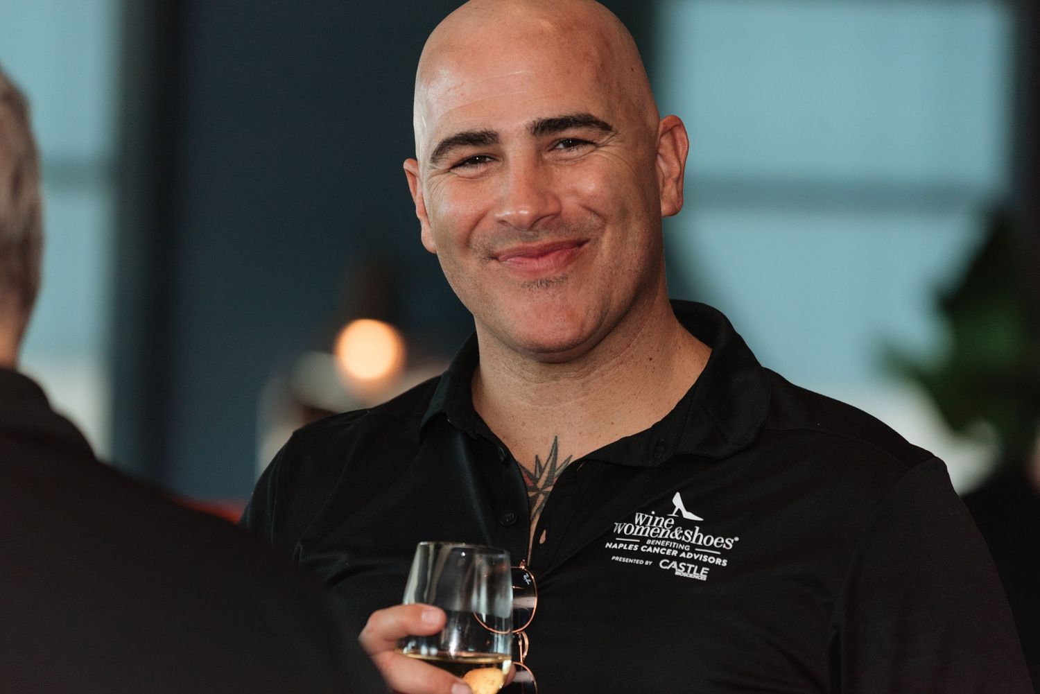 A person wearing a black chef's coat smiles while holding a wine glass in a restaurant setting.