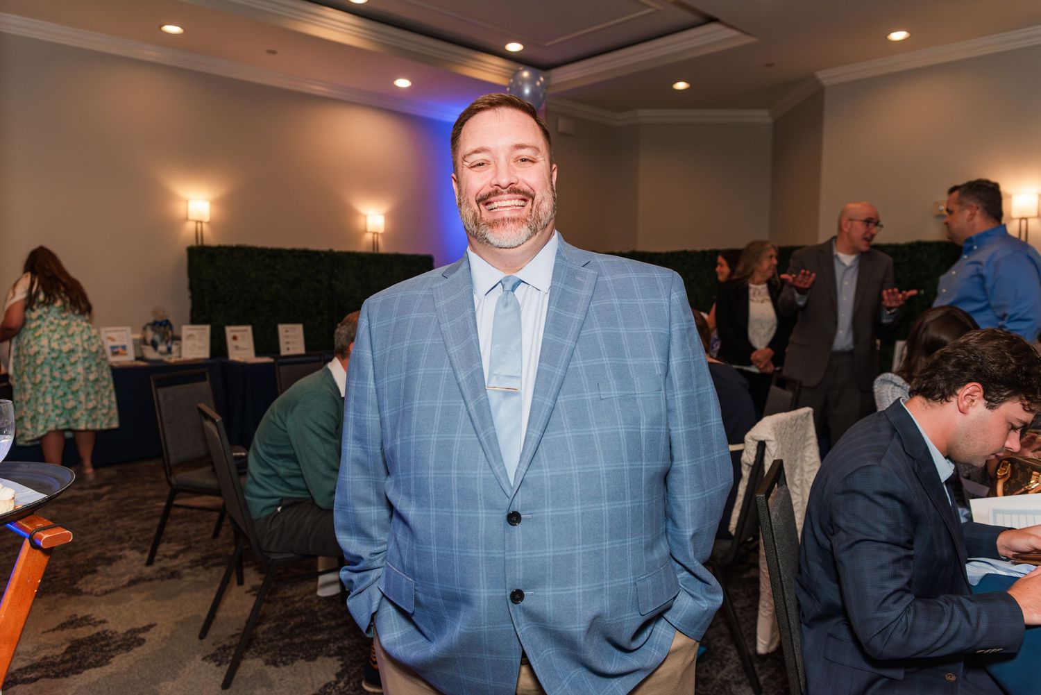 A person in a light blue suit and tie stands smiling in a banquet room with other guests seated at tables.