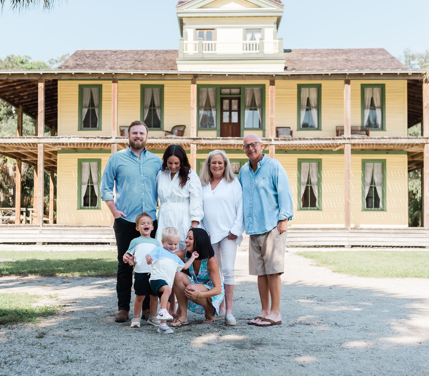 A group poses together in front of a grand yellow historic mansion with white trim and columns on a sunny day.