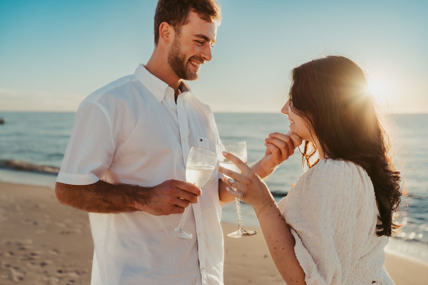 Couple enjoying wine glasses together on a sunny beach at sunset with golden light streaming behind them.