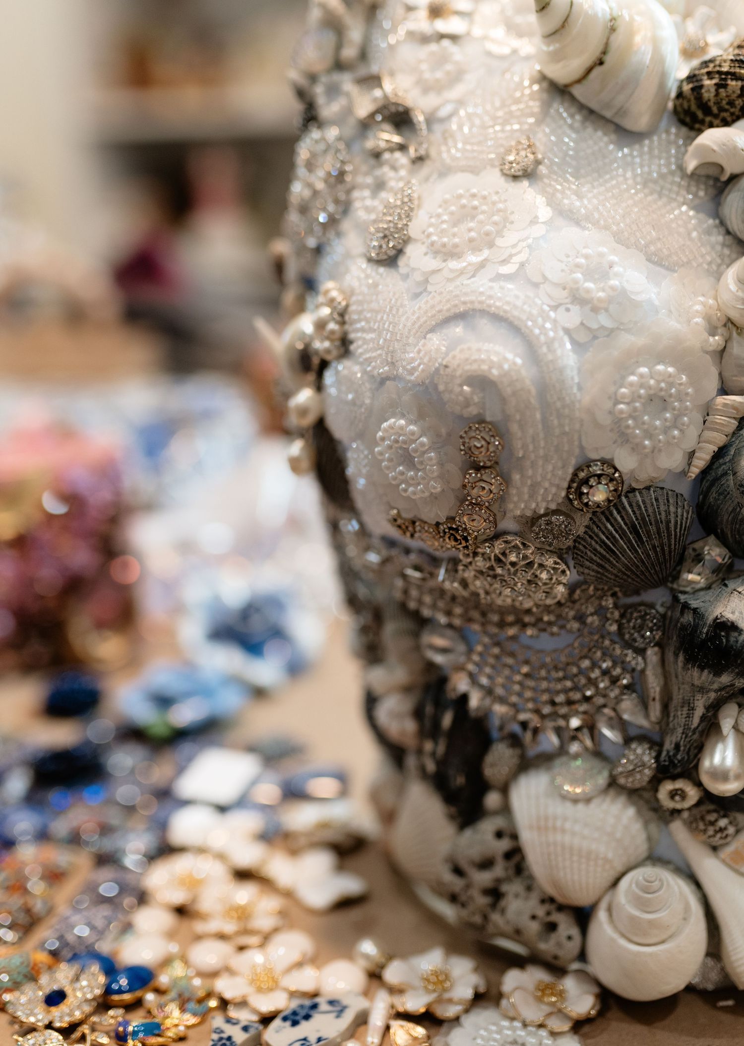 Close-up of an ornate seashell and pearl encrusted decorative vase on a craft table with scattered beads.